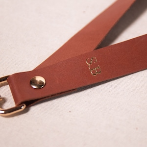 May include: A close-up of a brown leather lanyard with gold-coloured hardware. The lanyard has the initials "S.B." in gold lettering. The hardware includes a swivel clasp and a rivet.