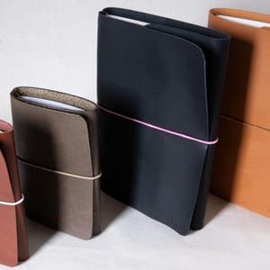 Puede incluir: Cuatro diarios de cuero en varios tamaños y colores. Los diarios son en tonos marrón, negro y beige. Cada diario está asegurado con una banda elástica. Los diarios están apilados uno al lado del otro.