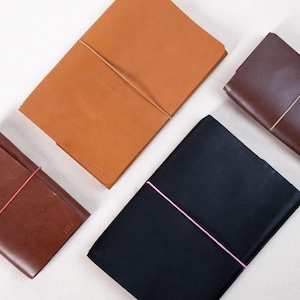 May include: Four leather journals in tan, dark brown, black, and medium brown. Each journal is rectangular and secured with an elastic band. The journals are arranged on a white surface.