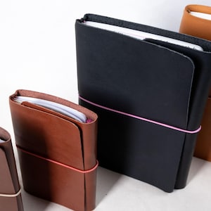 Pode incluir: Uma coleção de cadernos de couro em vários tons de castanho e preto. Cada caderno é preso com uma faixa elástica colorida. Os cadernos estão abertos para revelar papel branco no interior. Os cadernos são de tamanhos diferentes.