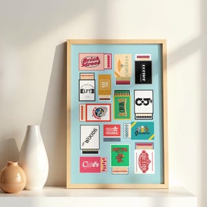 May include: A framed print featuring a collection of vintage matchbox designs. The matchboxes display various graphics and text, including "Brick Street," "The Woods," and "CALENDULA." The artwork is set against a light blue background.