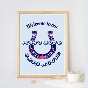 Puede incluir: Una impresión enmarcada azul claro con el texto "Welcome to our Mojo Dojo Casa House" en una fuente azul marino. Una herradura con una estrella rosa en el centro se representa en el medio de la impresión.