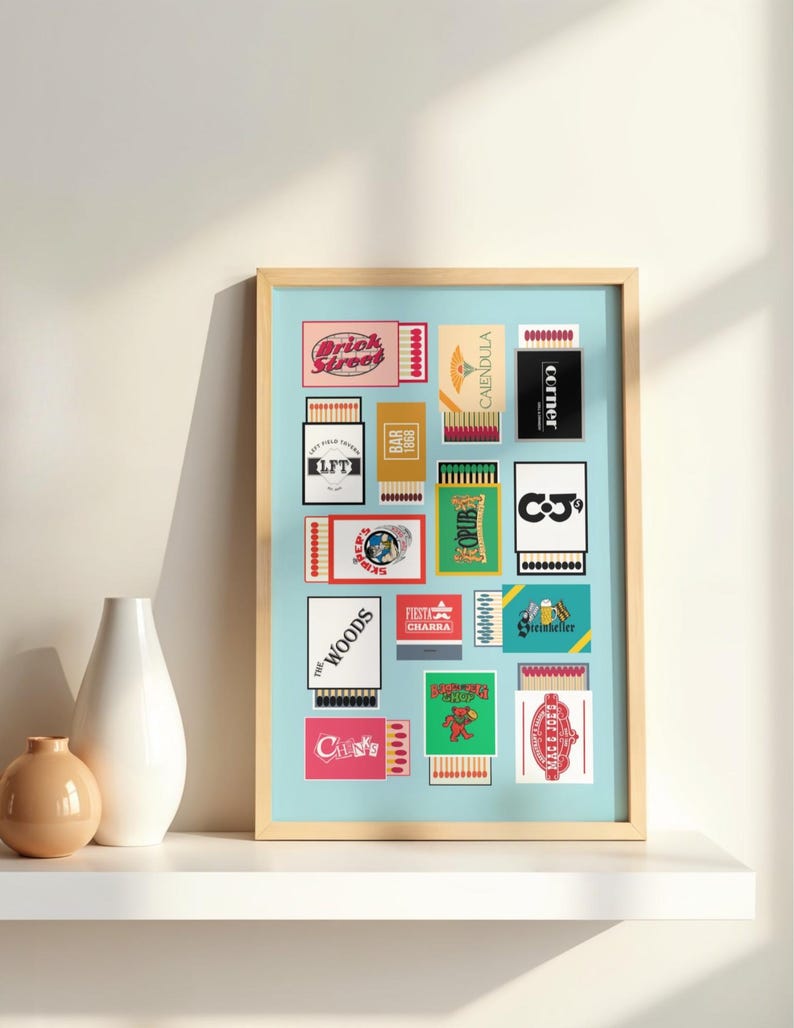 May include: A framed print featuring a collection of vintage matchbox designs. The matchboxes display various graphics and text, including "Brick Street," "The Woods," and "CALENDULA." The artwork is set against a light blue background.