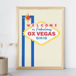May include: A framed print with a yellow, red, and blue Vegas-style sign that reads "Welcome to Fabulous Ox Vegas Ohio".