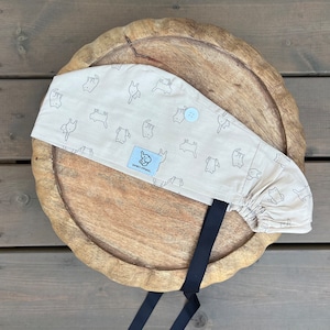 Op de afbeelding: Een beige scrubmuts met een zwarte stropdas en een witte knoop. De muts heeft een herhalend patroon van zwarte katten. De tekst "JUMBO CREW" is op de muts gedrukt.