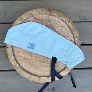 May include: White scrub hat with a pink heart pattern and a black tie. The hat has a button on the top and a small label with the text "Simply Inspired".