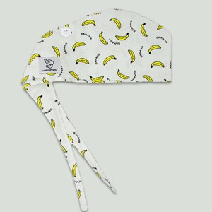 Pode incluir: Um gorro cir&uacute;rgico de tecido branco com um la&ccedil;o na parte de tr&aacute;s e um fecho de bot&atilde;o. O gorro tem um padr&atilde;o repetitivo de bananas amarelas com a palavra "banana" escrita em cada banana.