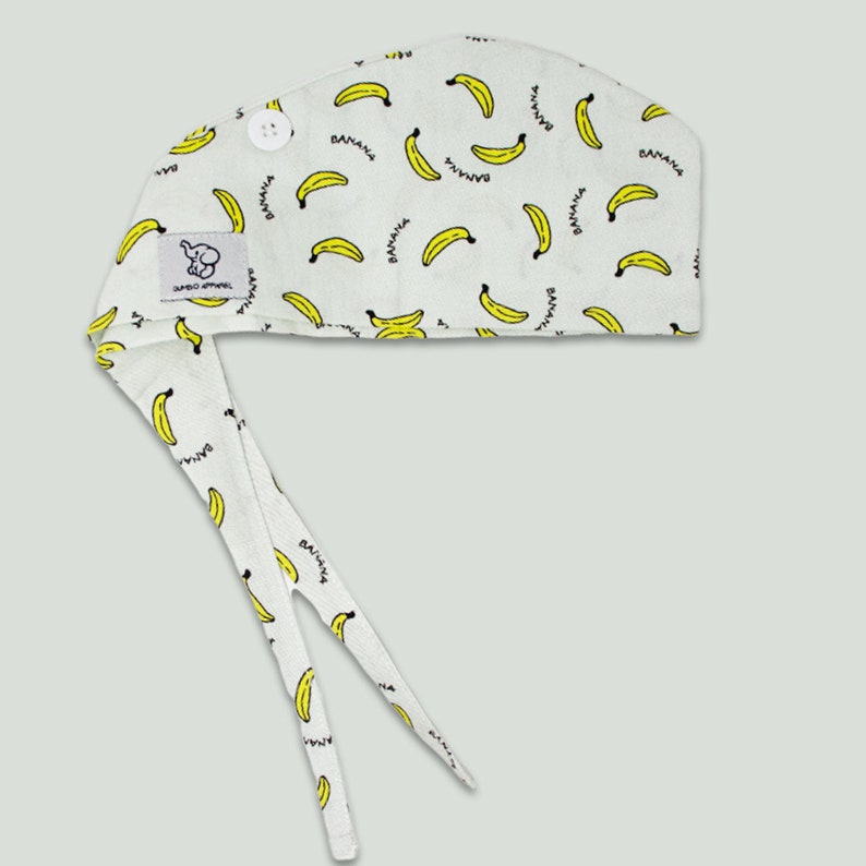 Pode incluir: Um gorro cir&uacute;rgico de tecido branco com um la&ccedil;o na parte de tr&aacute;s e um fecho de bot&atilde;o. O gorro tem um padr&atilde;o repetitivo de bananas amarelas com a palavra "banana" escrita em cada banana.