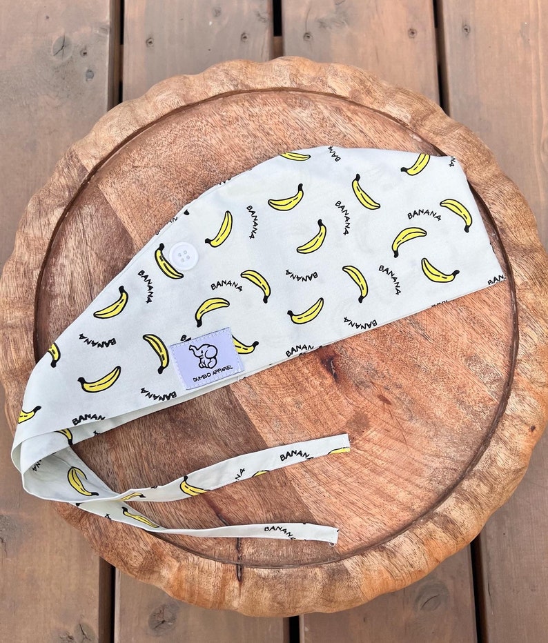 Pode incluir: Uma faixa de cabe&ccedil;a branca com um padr&atilde;o de banana amarela e o texto "BANANA" repetido por toda a superf&iacute;cie. A faixa de cabe&ccedil;a tem um pequeno logotipo de elefante com o texto "DUMBO APPAREL".