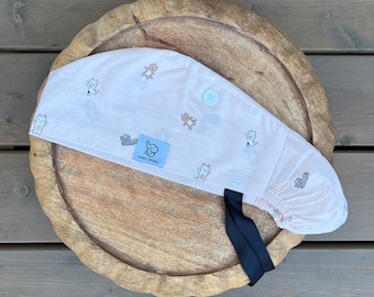 Puede incluir: Un gorro quirúrgico rosa claro con un estampado de gatos de dibujos animados. El gorro tiene un botón azul claro y una cinta negra. Una etiqueta azul con el texto "DUMBO APPAREL" está cosida en la parte delantera.