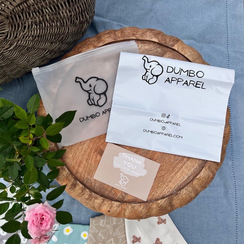 Pode incluir: Uma sacola de pl&aacute;stico branca com o texto "Dumbo Apparel" e uma pequena ilustra&ccedil;&atilde;o de elefante. A sacola est&aacute; sobre uma superf&iacute;cie de madeira com um cart&atilde;o de agradecimento e uma pequena planta.