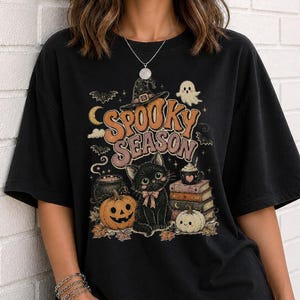May include: A black t-shirt featuring a graphic print with the text "SPOOKY SEASON" in orange and pink letters. The design includes a black cat with a pink bow, a jack-o'-lantern, a stack of books, a ghost, a cauldron, and a witch hat.