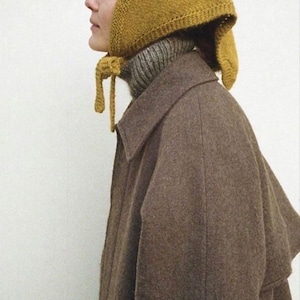 May include: A woman wearing a brown coat and a yellow knitted hat with a long tie. The hat has a pointed shape and covers the woman's ears.