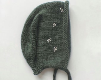 May include: A dark green knitted baby bonnet with white embroidered stars. The bonnet has ties to secure it under the chin.