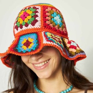 May include: A colorful, handmade crochet bucket hat. The hat features a patchwork design with squares of various colors, including orange, red, yellow, green, and blue. The brim is slightly flared, and the hat appears to be worn by a person.