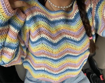 Op de afbeelding: Een kleurrijke, handgemaakte gehaakte trui met een zigzagpatroon. De trui heeft een mix van pastelkleuren, waaronder blauw, geel, roze en lavendel. De mouwen zijn lang en de halslijn is breed.