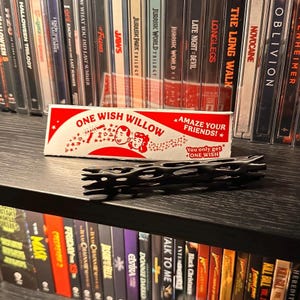 May include: A rectangular white cardboard box labeled ONE WISH WILLOW with red text and illustrations of two faces, positioned behind a black cylindrical object with a hollow, lattice-like structure featuring multiple irregular openings.