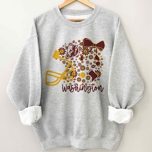 May include: A light grey crewneck sweatshirt featuring a graphic print of a yellow football helmet, a maroon bow, a bison head, and small flowers in shades of yellow and maroon. The word Washington is printed in maroon script below the design.