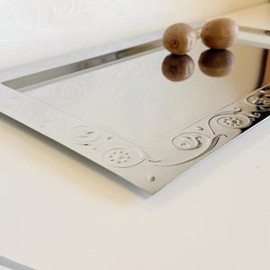 Puede incluir: Una bandeja de servicio rectangular de metal plateado con una superficie de espejo altamente reflectante. El borde ancho presenta un patrón de volutas en relieve con motivos florales circulares. Dos kiwis marrones descansan sobre la superficie.
