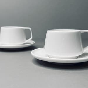 May include: Two white ceramic coffee cups with matching saucers. The cups have a modern, minimalist design with a slightly flared shape.