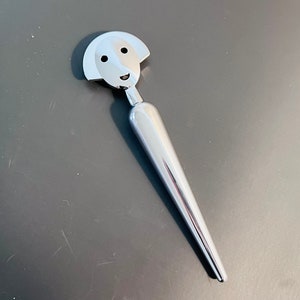 Può includere: Un utensile in metallo argentato con un disegno di un viso sulla parte superiore. L'utensile ha la forma di un cono lungo e sottile.