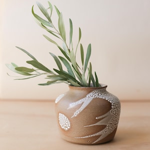 May include: A brown ceramic vase with white speckled patterns and a small opening, holding green leafy branches. The vase has a rounded shape and sits on a light brown surface. The leaves are long and slender.