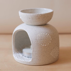May include: A speckled white ceramic essential oil diffuser. It features a round bowl and a cavity for a tea light. The diffuser has textured, raised dot patterns on the front and bowl, creating a handcrafted look.