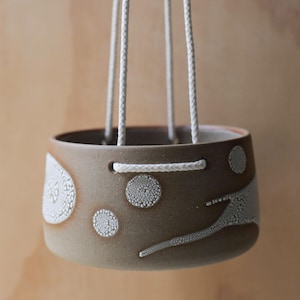 May include: A brown ceramic hanging planter with white textured patterns. The round planter is suspended by four white rope cords. The design features circular and linear patterns. The background is a light brown colour.