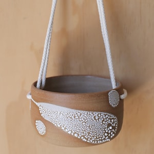 May include: A brown ceramic hanging planter with a white textured design. The planter is suspended by a white rope. The design features a large, abstract shape and two smaller circular shapes. The planter is designed for indoor or outdoor use.