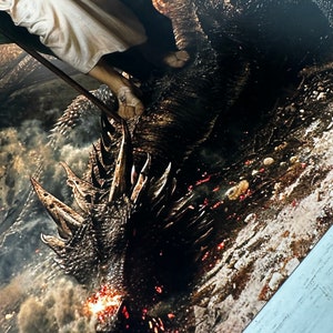 May include: A close-up of a dragon's head with flames coming out of its mouth. The dragon is lying on the ground and a person is standing on its back.