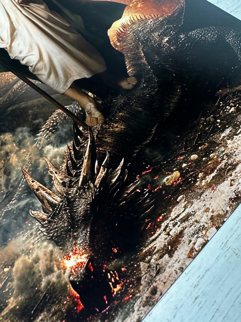 May include: A close-up of a dragon's head with flames coming out of its mouth. The dragon is lying on the ground and a person is standing on its back.