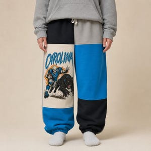 May include: Color-blocked sweatpants featuring a cream-colored panel on the left leg with a graphic print of a skeleton football player and a black panther, with the word CAROLINA in blue text above. The pants include black and blue panels.