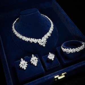 May include: A complete jewelry set featuring a necklace, bracelet, earrings, and ring. The set is adorned with clear, sparkling stones, arranged in a floral pattern. The jewelry is displayed in a dark blue velvet box, creating a luxurious presentation.