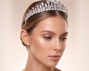 May include: A silver-toned tiara featuring a dense arrangement of clear, faceted crystals in various shapes, including pear and round cuts, set in a metallic band that arches across the top of the head to create a sparkling, elevated crown effect.