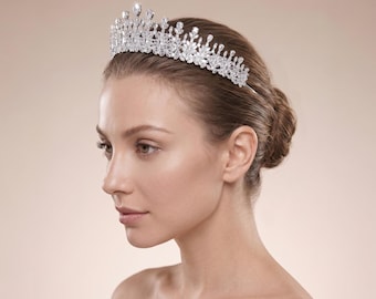 May include: A silver-toned tiara featuring a symmetrical arrangement of clear, faceted crystals in various shapes, including pear and round cuts, set in a metallic frame that arches across the top of the head with a pointed, elevated center design.