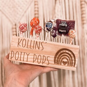 May include: A wooden sign with the text "KOLLINS POTTY PODS" and a rainbow with a heart in the center. The sign is holding a variety of lollipops with different flavors and colors.