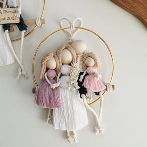 Op de afbeelding: Een gouden hoepel met een hartvormige knoop aan de bovenkant houdt een gezin van vijf garenpoppen vast. De poppen zijn gemaakt van wit en roze garen en hebben houten hoofden. De poppen hangen met touwtjes aan de hoepel. Het bordje staat "& Thomas Just 2022".