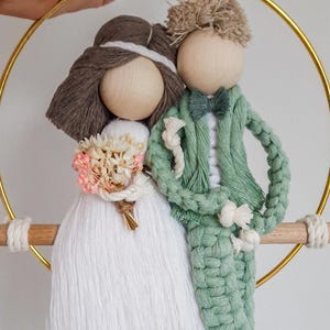 Può includere: Decorazione nuziale in macramè raffigurante una sposa e uno sposo. La sposa indossa un abito bianco e tiene un bouquet. Lo sposo è in abito verde con papillon. Le figure sono attaccate a un cerchio dorato con un tassello di legno.