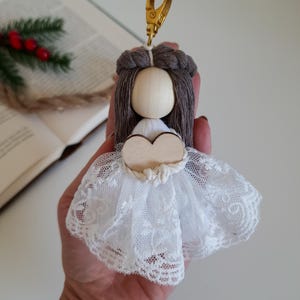 Könnte beinhalten: Ein kleiner Holzpuppen-Schlüsselanhänger mit braunen Haaren, einem weißen Spitzenkleid und einem Holzhäuschen. Die Puppe ist an einem goldenen Schlüsselring befestigt.