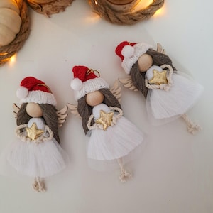 Può includere: Tre decorazioni natalizie a forma di angelo fatte a mano. Ogni angelo ha una testa di legno, capelli di filo marrone e ali di legno. Indossano abiti bianchi, dettagli a stella dorata e cappelli di Babbo Natale rossi e bianchi. Gli angeli sono esposti su una superficie bianca.