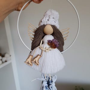 May include: A handcrafted angel decoration featuring a white knitted hat, brown yarn hair, and wooden wings. The angel is dressed in a white dress and holds a small posy. Wooden ice skates are suspended below. The ornament hangs from a white metal hoop.