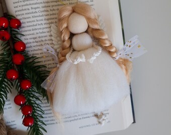 Puede incluir: Un adorno de ángel de fieltro blanco con pelo de lana rubio, falda de tul blanco y alas de purpurina dorada. El ángel está colgado de una cuerda dorada y descansa sobre un libro.