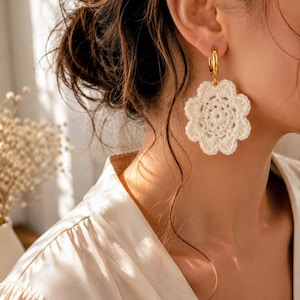May include: A gold hoop earring featuring a large, off-white crocheted flower pendant. The flower has eight rounded petals and a textured, circular center. The earring is suspended from a polished gold-tone metal hoop that secures to the ear.