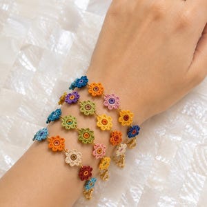 Pode incluir: Um conjunto de quatro pulseiras delicadas com uma corrente de pequenos motivos florais de crochê multicoloridos. Cada pulseira exibe uma sequência única de tons vibrantes, incluindo azul, laranja, rosa, amarelo, verde, vermelho e creme, conectados por elos de metal dourado finos e finalizados com um pequeno fecho lagosta.