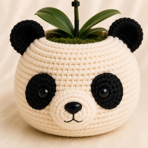 Pode incluir: Um vaso de crochê cor de creme em formato de rosto de panda, com orelhas pretas, manchas pretas ao redor de olhos brilhantes, um nariz de plástico preto e uma boca bordada, contendo uma planta verde com musgo.