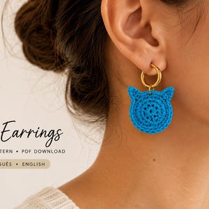 Pode incluir: Um brinco de crochê azul brilhante em forma de gato pendurado em uma argola de metal dourado. O texto na imagem diz: Cat Earrings, CROCHET PATTERN, PDF DOWNLOAD, PORTUGUÊS, ENGLISH. O brinco apresenta um design circular com duas orelhas.