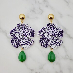 Pode incluir: Um par de brincos com um pino dourado, uma flor de crochê em fio branco e roxo e uma conta verde em forma de gota. O design floral consiste em cinco pétalas e a conta verde pende da parte inferior central.