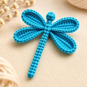 Pode incluir: Uma libélula azul brilhante feita de fio de crochê. O inseto apresenta um corpo longo e segmentado, uma pequena cabeça arredondada e quatro asas alongadas distintas com uma textura costurada visível em todo o design feito à mão.