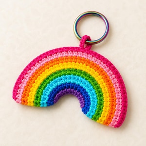 Pode incluir: Um porta-chaves de arco-íris em croché com arcos concêntricos de fio cor-de-rosa, laranja, amarelo, verde, azul e roxo. O arco-íris têxtil está preso a um anel metálico circular e iridescente que apresenta um espectro de cores metálicas.