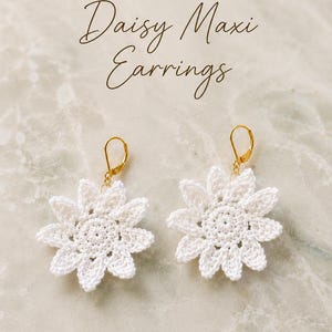 Pode incluir: Um par de brincos de crochê branco em forma de margarida com fechos de alavanca dourados. O texto MTOMCROCHE CROCHET DESIGNS Daisy Maxi Earrings é visível acima dos brincos. O design floral apresenta um padrão circular central.